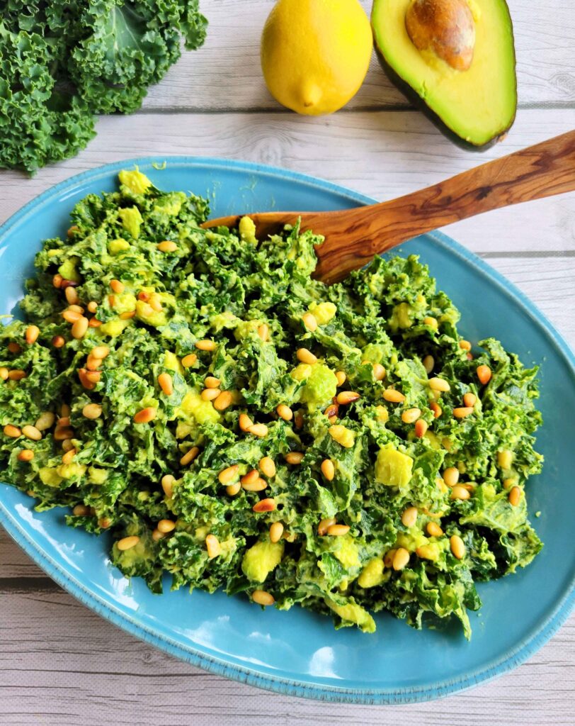 Fresh kale, lemon, and avocado next to a bowl of this gut-healthy, anti-inflammatory massaged kale salad with a wooden serving spoon