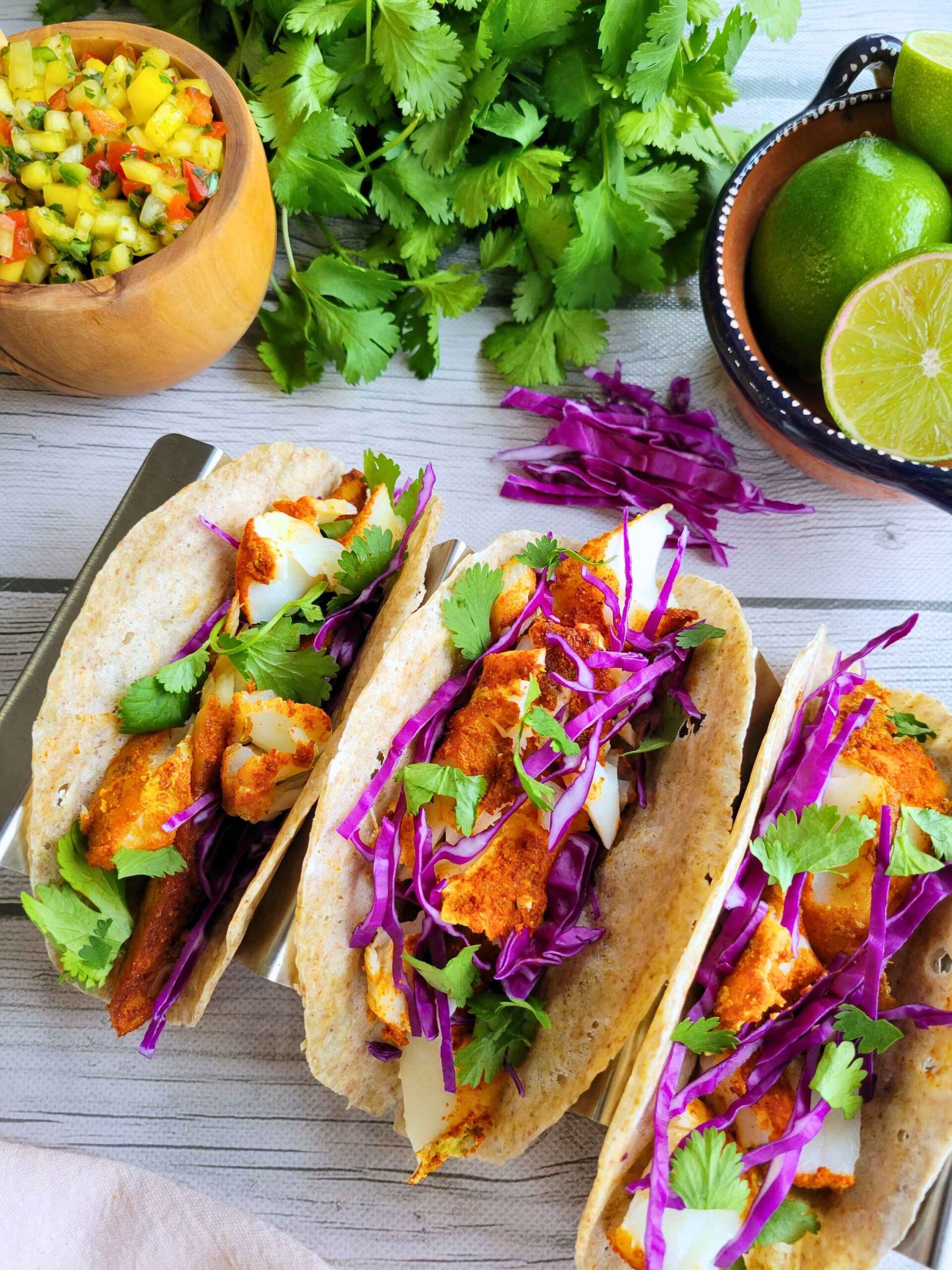 3 Baked anti-inflammatory fish tacos on sprouted buckwheat tortillas topped with purple cabbage, cilantro and next to a bowl of gut-healthy mango salsa, limes and fresh toppings.
